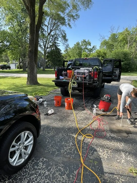 Woman cleaning car mat with mobile detailing equipment and truck on driveway.